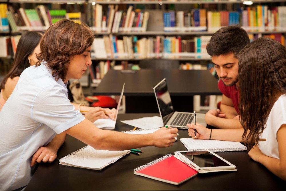 Grupo de estudiantes colaborando en una biblioteca con portátiles, libretas y tabletas mientras cursan posgrados en España, enfocados en un proyecto académico en equipo.