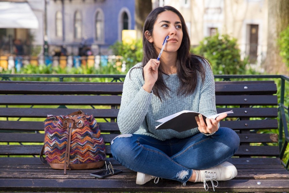 Joven sentada en un banco de parque con libreta en mano, pensando y planificando su formación profesional mientras considera estudiar posgrados en España.